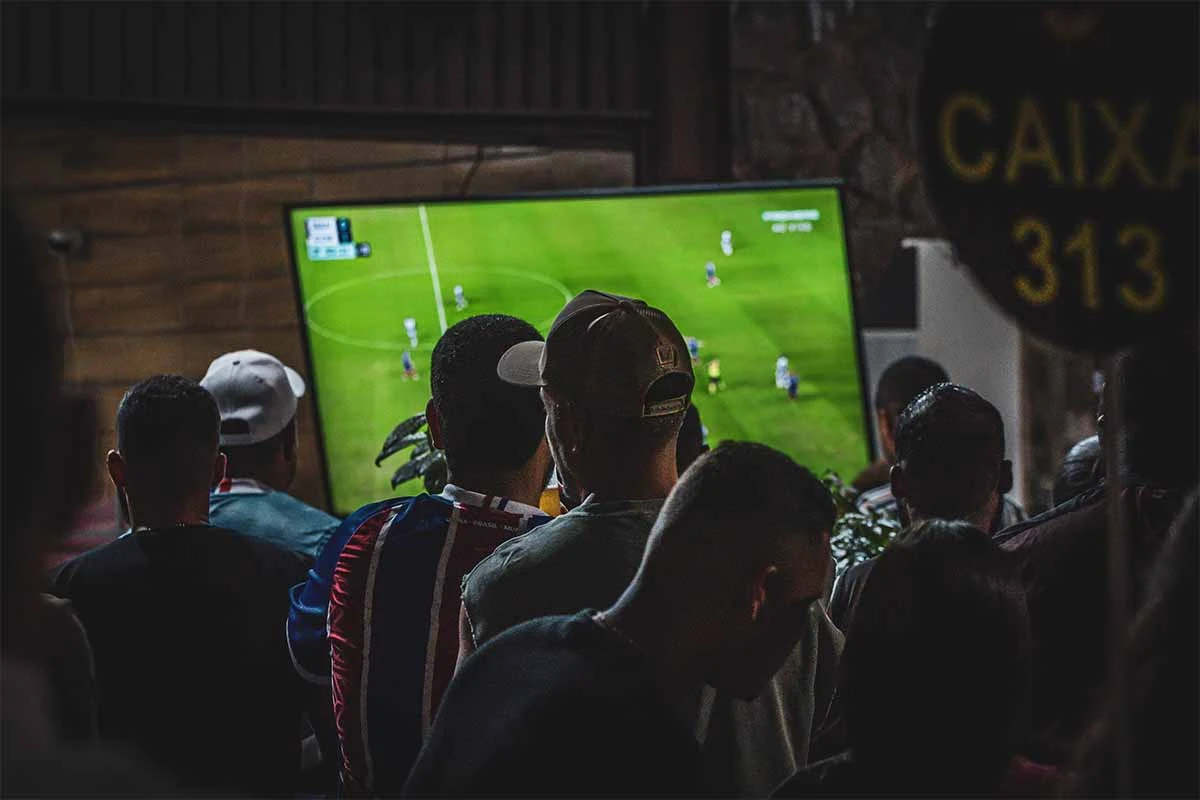 Grupo de personas viendo un partido de fútbol en pantalla grande dentro de un local cerrado.